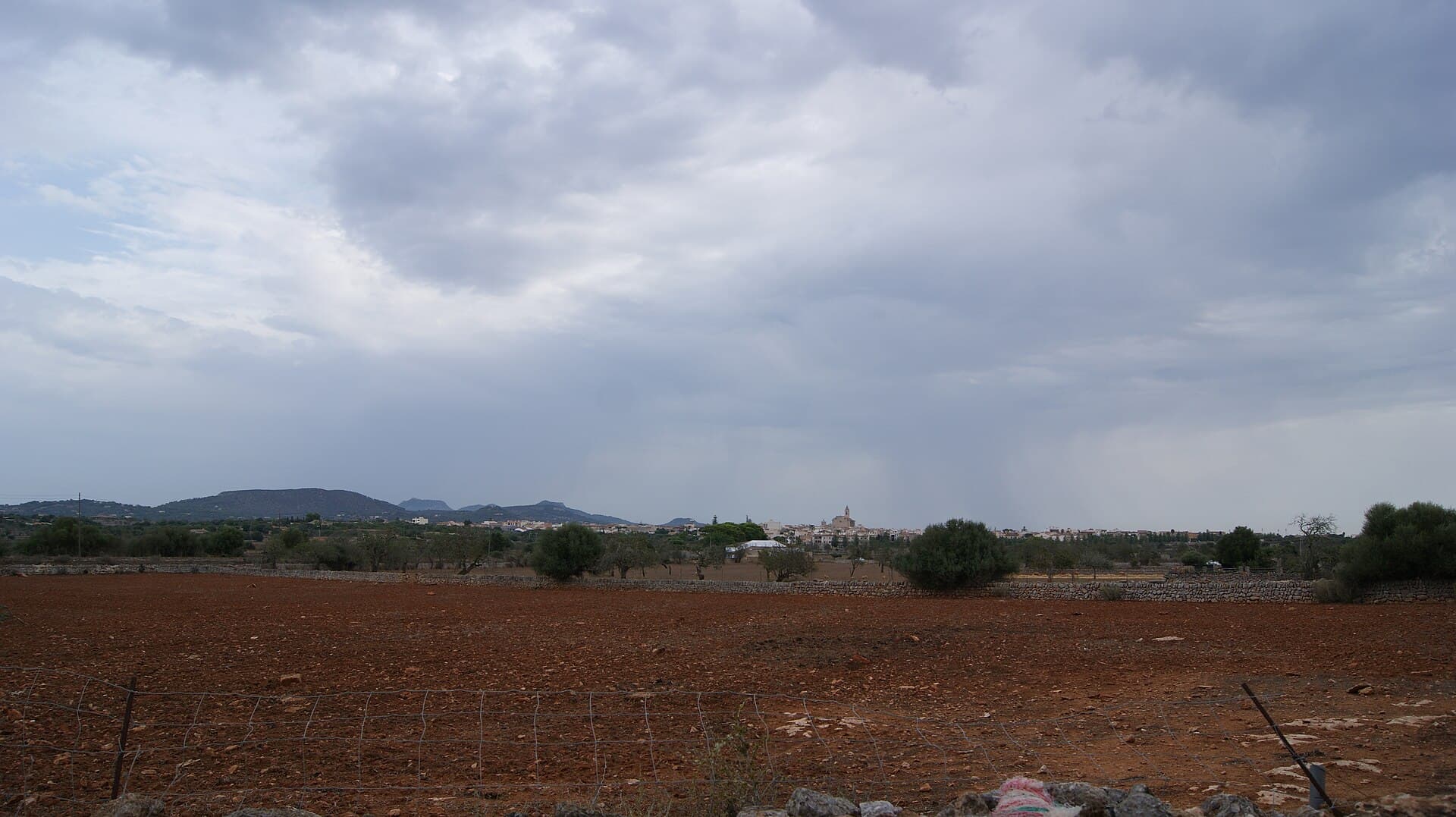 Ort Santanyí über Terra-rossa-Feld und Trockenmauer unter bewölktem Himmel, Mallorca